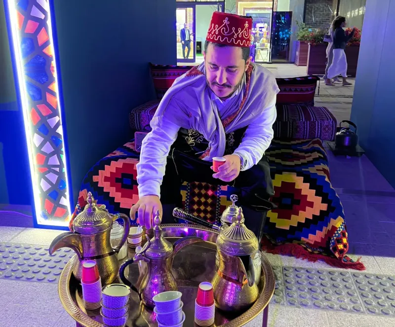 Man in a traditional hat preparing Arabic coffee on a colourful majlis rug