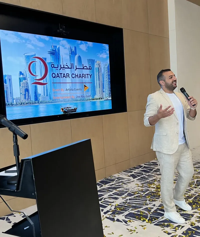 Host in a white suit addressing guests beside a Qatar Charity presentation screen