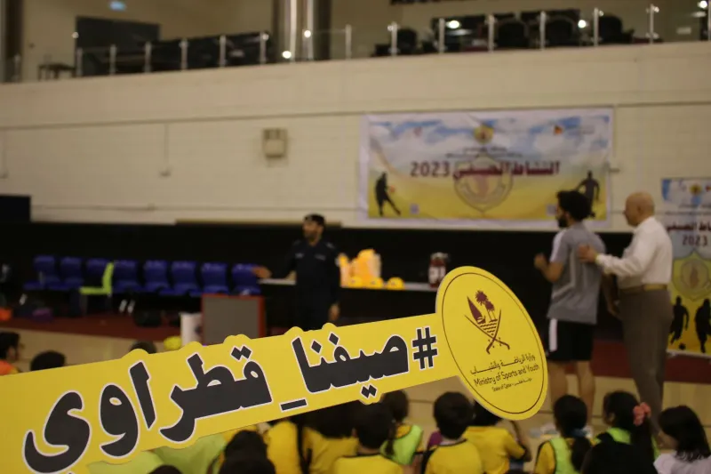 Children in a sports hall holding a giant yellow Qatari summer camp sign