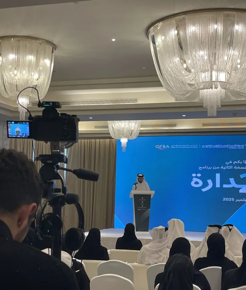 Speaker presenting at a podium on stage with Sadara 2 branding and a camera crew