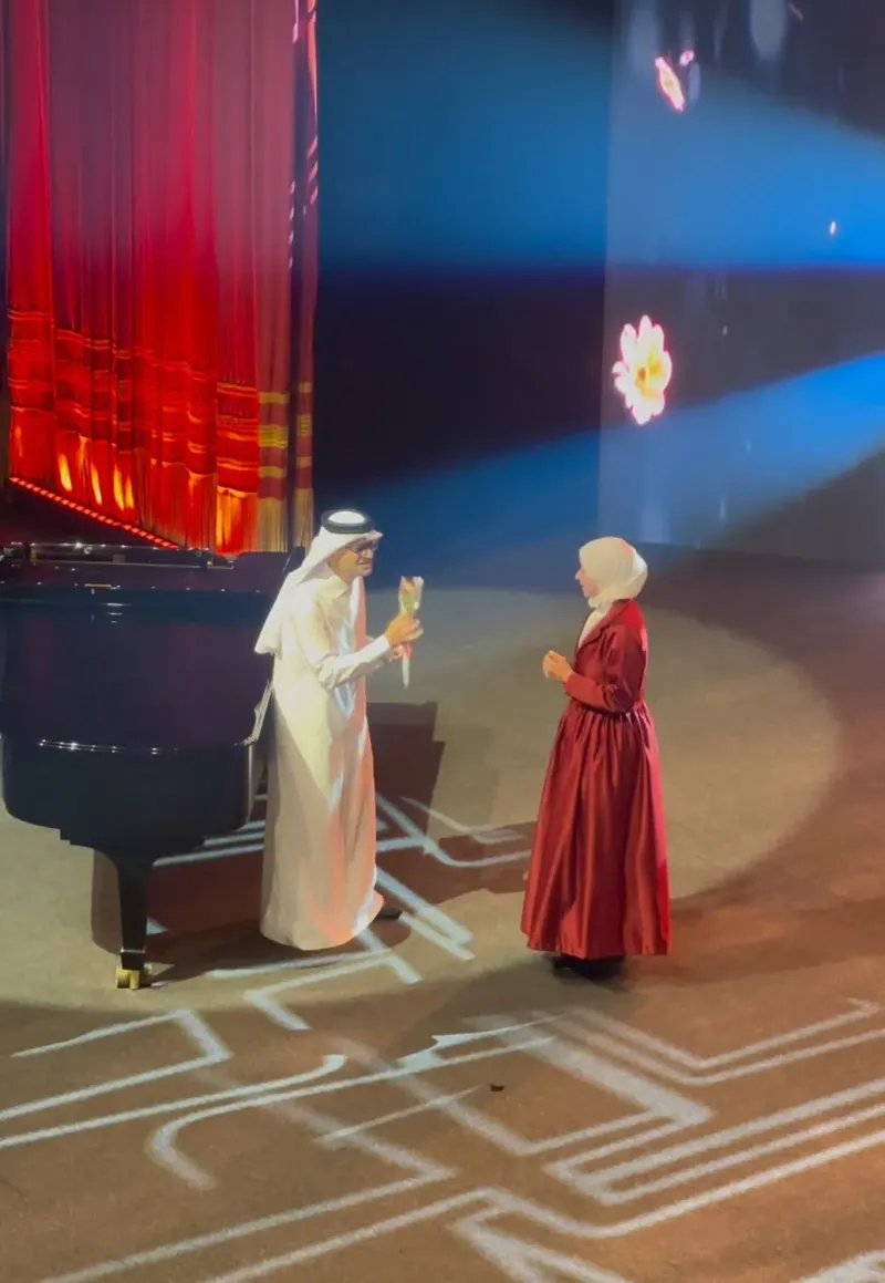 Two performers on stage beside a grand piano under blue lighting