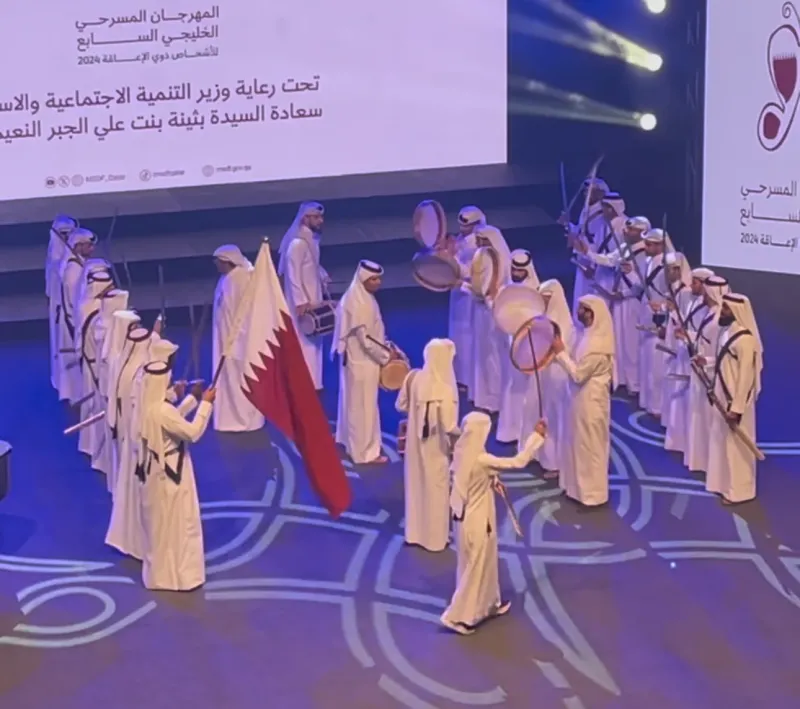 Gulf theatre performers in white thobes dancing on stage with a Qatari flag