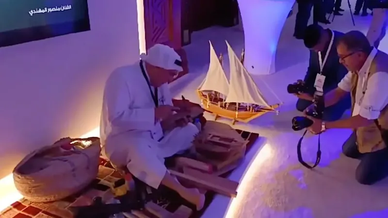 Craftsman building a wooden dhow model while a photographer captures the moment