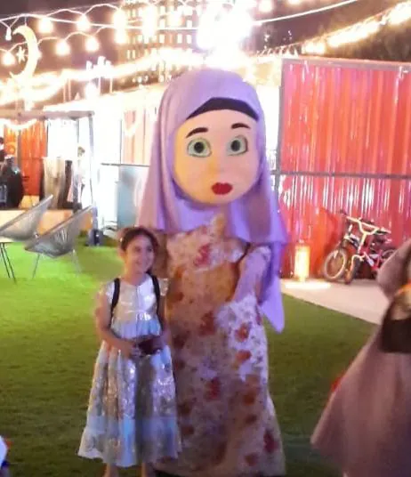 Girl in a floral dress posing beside a large costumed hijab mascot at night
