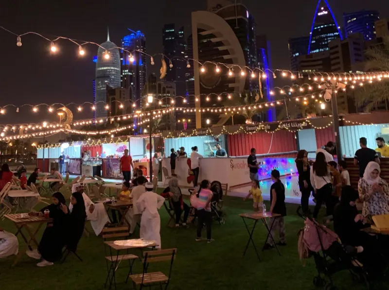Guests at lawn tables under string lights beside illuminated festival cabins at night